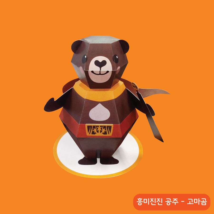 노란 테두리 원형 받침 위에 선 진갈색 고마곰 페이퍼토이, 붉은 망토와 무늬 허리띠에 배에는 흰 밤톨 무늬가 있다
