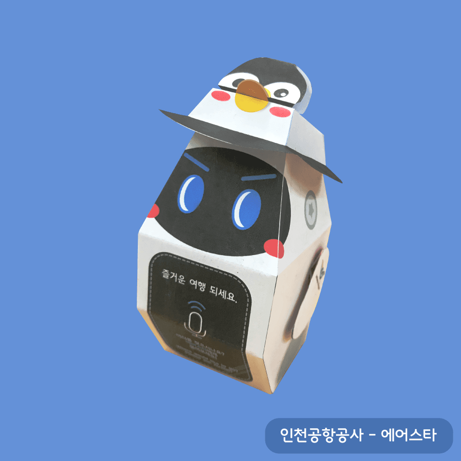 파란 눈의 안내 로봇 에어스타 위에 펭귄이 얹힌 페이퍼토이, 몸통에 즐거운 여행 되세요 문구와 마이크 아이콘이 있다